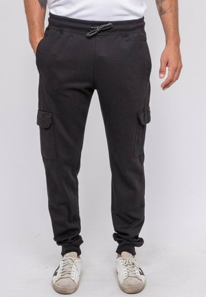 Homme portant un pantalon cargo jogger noir avec poches latérales, cordon blanc, t-shirt blanc et baskets blanches, debout devant un fond uni.