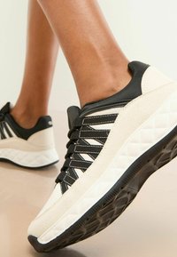 Sneaker negru și crem cu un exterior texturat, șireturi negre și o talpă groasă, cu un model. Designul prezintă accente geometrice și un toc căptușit.