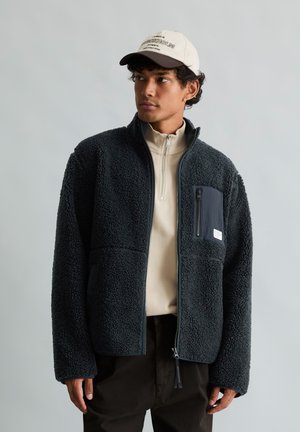Ung man som bär en svart fleecejacka med en dragkedjeficka och en beige tröja med kvartslång dragkedja, kombinerat med en beige och svart basebollkeps.