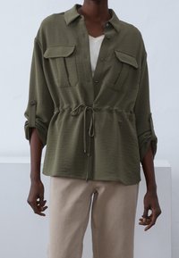 Olive green shirt with chest pockets, button closure, and adjustable drawstring waist. Soft texture, relaxed fit, and rolled sleeve cuffs.