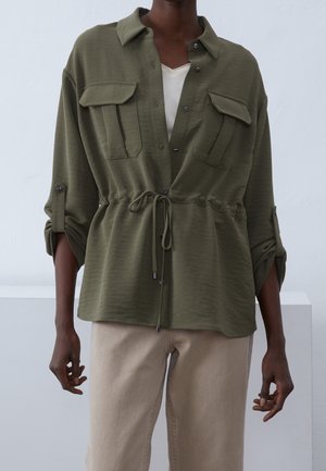 Chemise vert olive avec poches poitrine, fermeture à boutons, et taille ajustable avec cordon. Texture douce, coupe décontractée et poignets de manches retroussés.