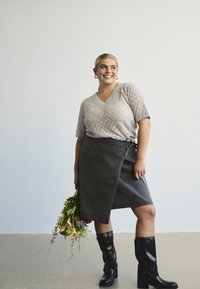 Naeratav naine hoiab lilledega, kandes heledat kudumit, tumedat salli-seelikut ja musti põlvikute kõrgusi saapaid, seistes ühtlase tausta ees.