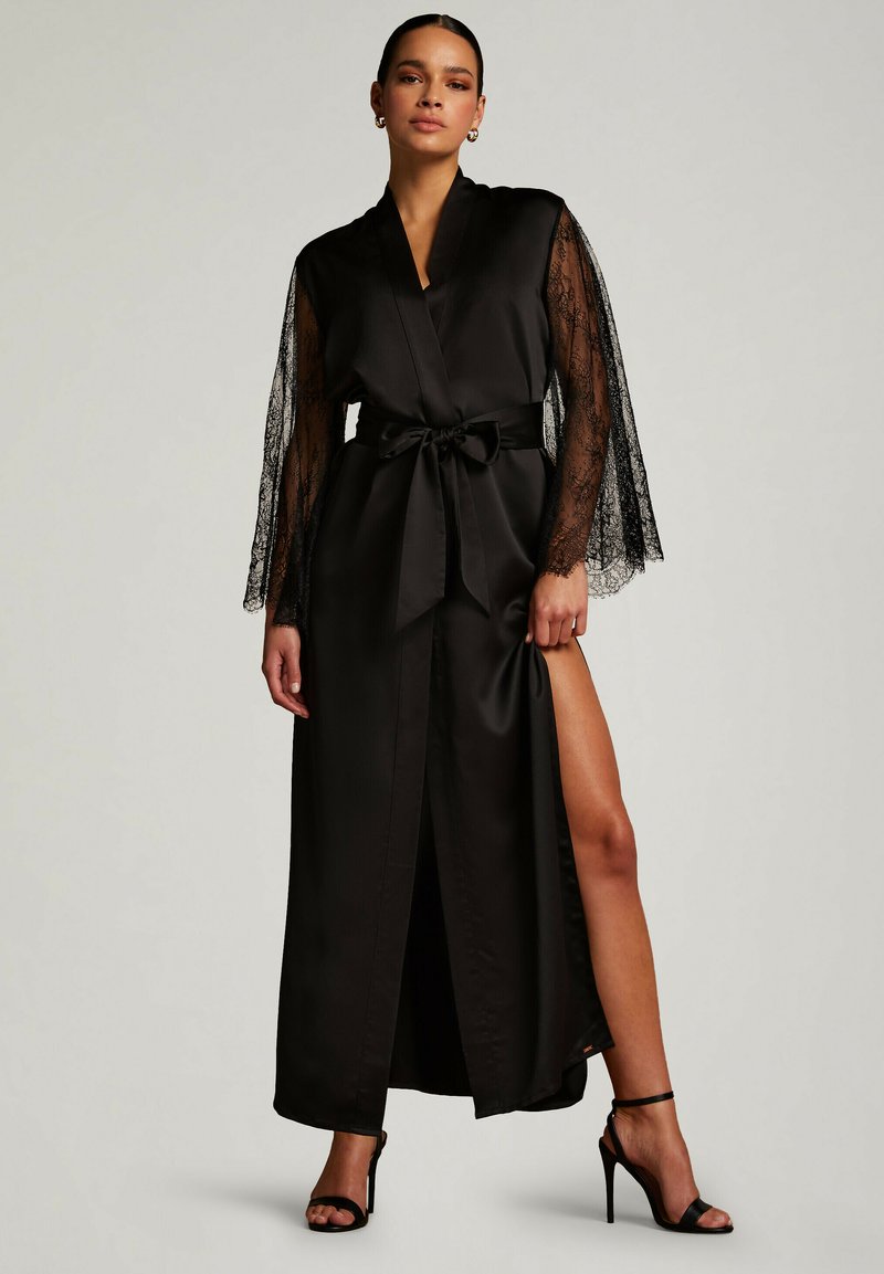 Robe di satin nero con maniche in pizzo, design avvolgente, cintura da annodare in vita, spacco laterale che mette in mostra la gamba, indossata con sandali neri con tacco alto.