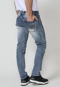Jeans de denim azul desgastados con un corte ajustado, cinco bolsillos, detalles de costura y acentos de logo en la parte trasera; combinados con zapatillas negras.