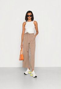 Calças bege de cintura alta com cinto a atar, top branco, mala laranja e ténis brancos com detalhes em néon. Modelo a usar óculos de sol.