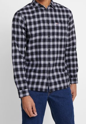 Mies, joka pukeutunut mustavalkoruudulliseen pitkähihainen nappipaitaan, jossa on pieni logo rinnassa, ja sinisiin farkkuihin.