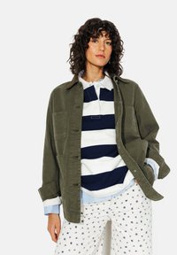 Veste olive avec poches avant, portée par-dessus un polo rayé bleu marine et blanc, superposée sur une chemise bleu clair et un pantalon blanc à motifs.