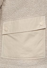 Beige fleece materiaal met een gestructureerd oppervlak, voorzien van een roomwitte katoenen zak die met twee ronde knopen is bevestigd.