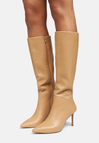 Botas de cuero beige hasta la rodilla con punta afilada y tacón de aguja de 10 cm, que cuentan con un cierre lateral y una textura suave y brillante.