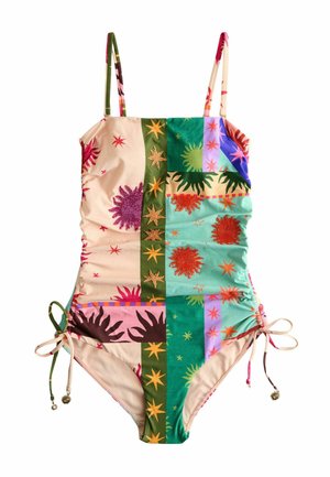 Kobieta z kręconymi włosami ubrana w bezramienny kostium kąpielowy w stylu patchwork, stoi na piaszczystej plaży, trzymając plecioną torbę.