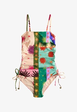 Femme aux cheveux bouclés portant un maillot de bain bustier à motif patchwork, debout sur une plage de sable, tenant un sac tissé.