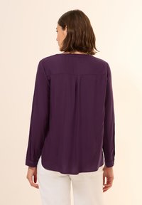 Blouse à manches longues, de couleur violet foncé, avec une coupe droite, un détail de pli au dos et des poignets boutonnés, fabriquée en tissu léger et lisse.