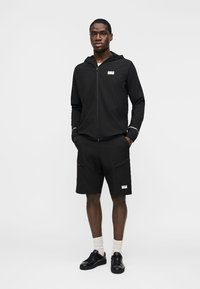 Άνδρας ντυμένος με μαύρο φούτερ με φερμουάρ και μαύρο σορτς, φορώντας λευκές κάλτσες και μαύρα αθλητικά παπούτσια, στέκεται με τα χέρια στις τσέπες.