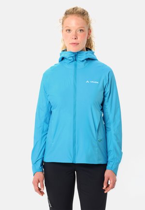 Junge Frau mit lockigem blondem Haar, die eine leuchtend blaue Kapuzenjacke und schwarze Hose trägt, vor einem schlichten Hintergrund stehend.