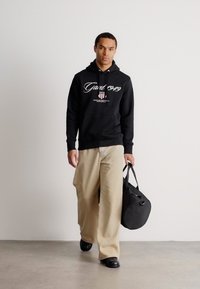 Bărbat care merge, purtând un hanorac negru "Gant 1949", pantaloni bej cu croială largă, pantofi negri, ținând în mâna dreaptă o geantă de duffle neagră cu curea.