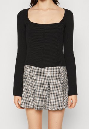 Femme portant un haut noir à manches longues côtelé avec un décolleté carré et une jupe plissée à carreaux beige et noire.