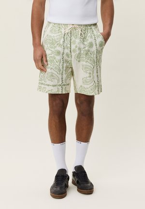 Man draagt witte korte broek met groene paisley-patronen, witte sokken en zwarte sneakers, staand met handen in de zakken.