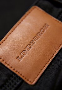 Patche en cuir marron avec le nom de la marque embossé "Lindbergh" cousu sur un tissu noir.