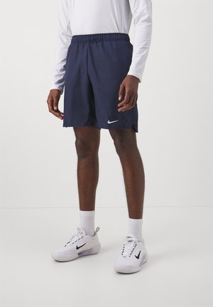 Man wearing navy athletic shorts, white long-sleeve shirt, white crew socks, and white Nike sneakers standing against a plain background.