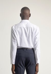 Camisa blanca de manga larga con cuello clásico, combinada con pantalones oscuros azules que tienen un cinturón negro texturizado y dos bolsillos traseros.