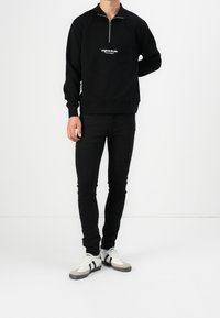 Czarna bluza rozpinana na zamek z białym logo na piersi, zestawiona z dopasowanymi czarnymi jeansami i czarno-białymi trampkami z paskowymi akcentami.