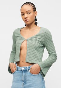 Νεαρή γυναίκα με πλεξουδωτά μαλλιά, φοράει cropped ζακέτα ανοιχτό πράσινο με υφή, κουμπωμένη στο πάνω μέρος, και ανοιχτό μπλε τζιν ψηλόμεσο, με τα χέρια στις τσέπες.