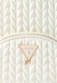 Borsa bianca trapuntata con una texture intrecciata, caratterizzata da una targhetta logo triangolare dorata con la scritta "GUESS U.S.A." e un dettaglio con cerniera dorata.