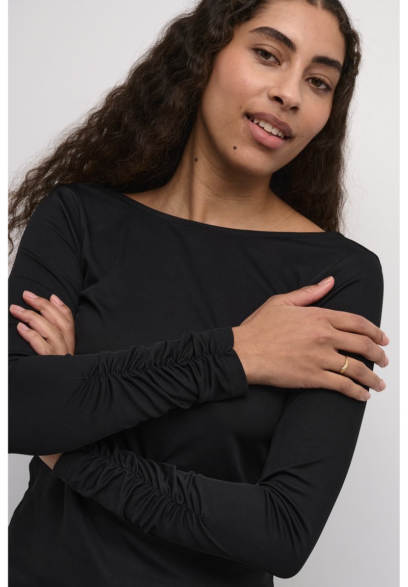 Schwarzes Langarmoberteil mit gerippter Textur, Bootsausschnitt und gerafften Akzenten an den Handgelenken. Die Arme des Models sind verschränkt.