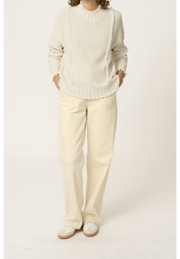 Person, der einen cremefarbenen Strickpullover, lockere beige Hose und weiße Sneaker mit Gummisohlen trägt, steht mit Händen in den Taschen.