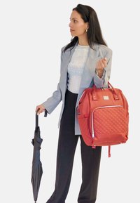 Mochila acolchada roja con correas para los hombros y un zipper frontal, sostenida por una persona con un blazer gris y pantalones negros, que sostiene un paraguas.