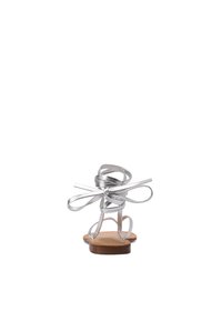 Sandalo argento con cinturini e suola piatta, caratterizzato da anelli allungati e fiocchi sul retro, poggiato su una soletta color beige.