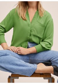 Femme portant un chemisier vert vif et un jean bleu clair assise en tailleur sur un banc en bois, chaussures noires visibles aux pieds.