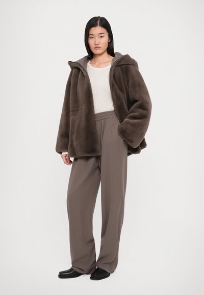 Kvinna klädd i en brun oversized fuskpälsjacka, beige topp, vida taupe byxor och svarta skor, stående mot en plain bakgrund.