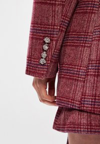 Plaid jacket in red and grey with blue accents. The sleeve features four silver leaf-shaped buttons and a textured fabric.