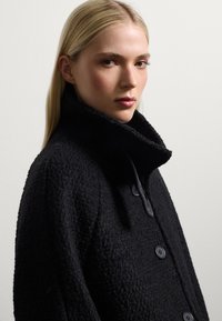 Cappotto nero textured con colletto alto, caratterizzato da grandi bottoni. Il materiale sembra morbido e tessuto, offrendo un design strutturato ma accogliente.