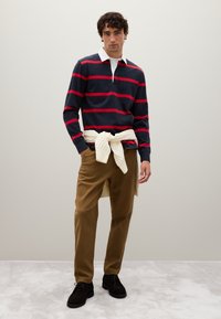 Uomo che indossa una maglietta da rugby blu navy con strisce rosse, pantaloni marroni, scarpe nere e un maglione di crema annodato in vita, in piedi.
