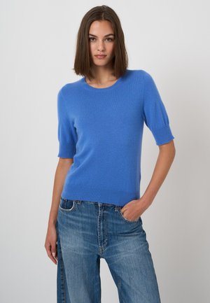 Young woman with straight brown hair wearing a blue short-sleeve sweater and blue jeans, standing with one hand in her pocket.