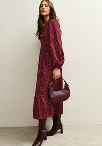Burgunderrotes Kleid mit weißen Polka-Dots; lange Puffärmel, gestufter Rock. Accessoire: kleine burgunderrote Handtasche. Paar braune Stiefeletten mit Absatz.