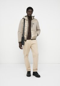 Beige gewatteerde jas met een rits, capuchon en één borstzak. Draagt over een donkerbruine top, gecombineerd met lichte broeken en zwarte laarzen.