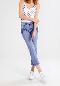 Débardeur blanc à pois noirs, jean taille haute bleu clair avec imprimé floral sur les jambes, associé à des baskets blanches.