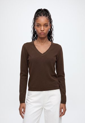Femme aux cheveux tressés portant des boucles d'oreilles en perles, un pull marron à col en V et manches longues, et un pantalon blanc, debout devant un fond blanc uni.