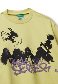 Gelbes Sweatshirt mit rundem Ausschnitt, das einen schwarzen und lila grafischen Druck zeigt, einschließlich Figuren und dem Text "THRILL SEEKER." Weiche Stofftextur.