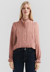 Blouse de couleur rose pâle avec un col haut, un devant plissé, des accents en dentelle et de longues manches bouffantes avec des poignets froncés. Assortie à un jean foncé.