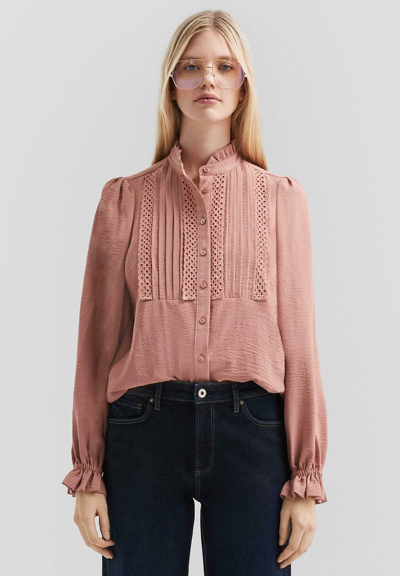 Blouse de couleur rose pâle avec un col haut, un devant plissé, des accents en dentelle et de longues manches bouffantes avec des poignets froncés. Assortie à un jean foncé.