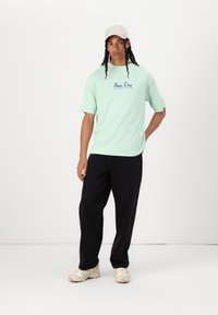 Mento grøn kortærmet t-shirt med blåt "New Era" logo. Bæret med løse sorte bukser og lyse sneakers. Enkel, afslappet pasform.