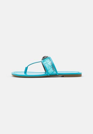 Flad turkisblå sandal med en tåcirkel og bred rem med tekstureret mønster og en dekorativ sølvemblem.