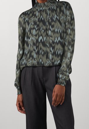 Vrouw draagt een blouse met hoge hals en lange mouwen met een abstract zwart, groen en lichtblauw patroon, ingestopt in donkergrijze wijde broek.