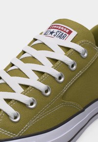 Gros plan sur une basket Converse All Star vert olive avec lacets blancs, œillets métalliques, coutures blanches et détail de semelle en caoutchouc.