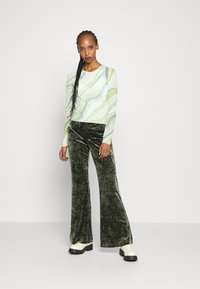 Blusa de manga longa com um padrão ondulado em verde e creme, combinada com calças de veludo verde e sapatos brancos chunky. Texturas suaves visíveis.