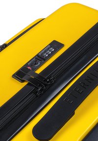 Valise rigide jaune avec fermetures éclair noires et verrouillage par combinaison, présentant une finition texturée lisse et un accent de marque sur le côté.